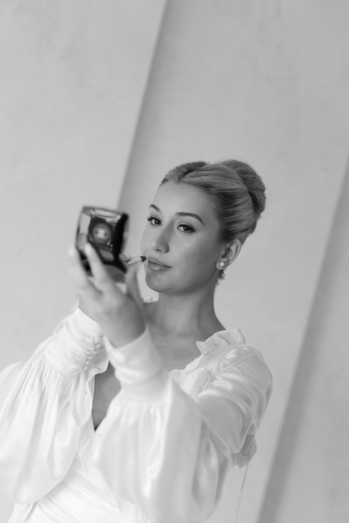 A woman in a white blouse applies lipstick while looking into a compact mirror—capturing a timeless, poised moment reminiscent of the glamour often seen at the Ariana and Julian Borghese wedding. The photo is black and white, and her hair is styled in an elegant updo.
