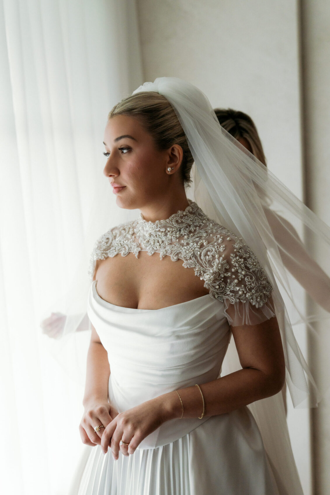 A bride in a white wedding dress with lace detailing and a veil stands by a window, looking outside with soft natural light illuminating her face, capturing the timeless elegance of the Ariana and Julian Borghese wedding.