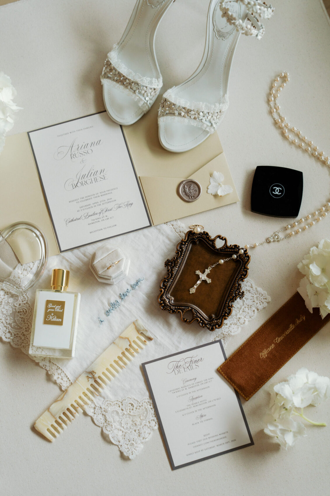 Flat lay of bridal accessories from the Ariana and Julian Borghese wedding, including white heels, invitations, perfume, pearl necklace, vintage coin, lace handkerchief, hair comb, Chanel box, rosary, and white flowers arranged on a neutral surface.