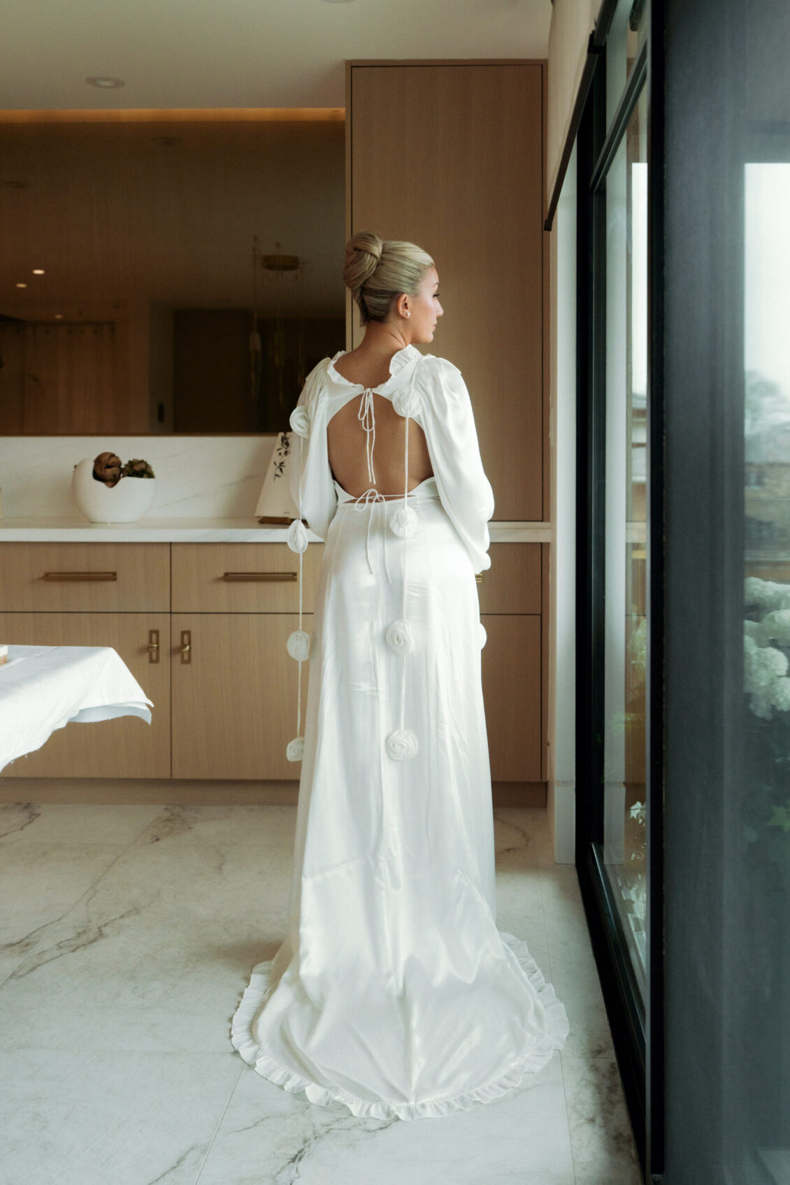A woman in a long white dress with an open back stands indoors by a large window, facing away—evoking the elegant ambiance of the Ariana and Julian Borghese wedding. Her hair is in a neat bun, and the room features marble floors and light-colored cabinets.