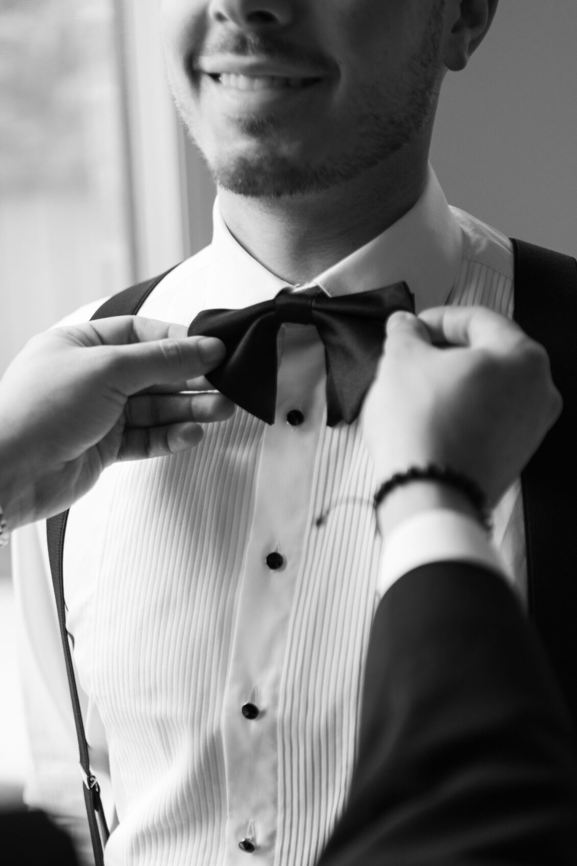 A person adjusts a bow tie on a smiling man wearing a dress shirt and suspenders at the Ariana and Julian Borghese wedding. The image is in black and white and shows only the upper body.