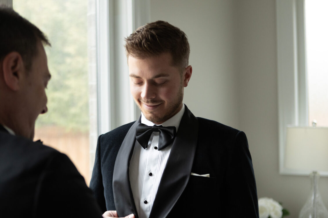 A man in a black tuxedo and bow tie stands indoors, smiling and looking down while another person faces him. Soft natural light comes through a window in the background, capturing a candid moment from the Ariana and Julian Borghese wedding.