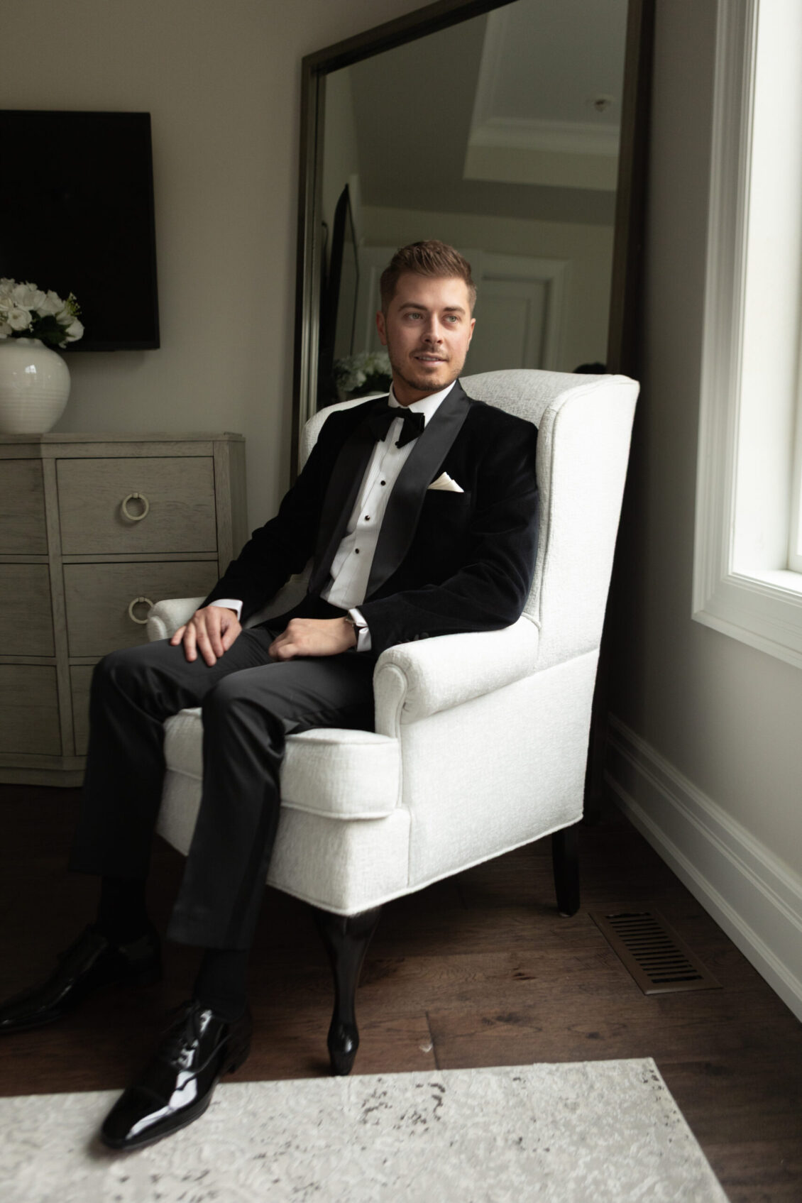 A man in a black tuxedo sits on a white armchair in a bright room, with a dresser and large mirror behind him, capturing the elegant atmosphere of the Ariana and Julian Borghese wedding.