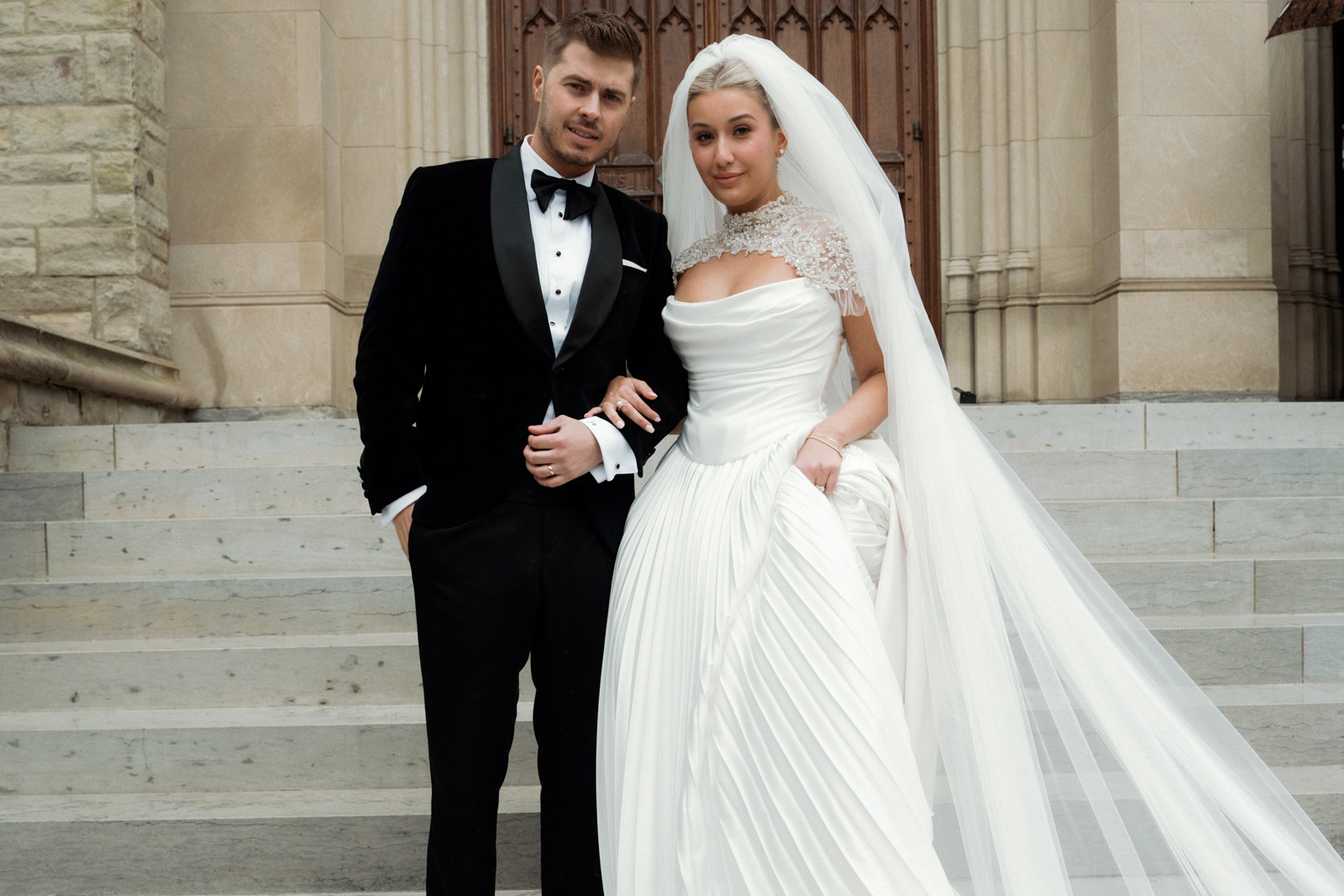 A bride in a white gown and veil stands arm-in-arm with a groom in a black tuxedo on stone church steps, posing for a wedding photo at the Ariana and Julian Borghese wedding.