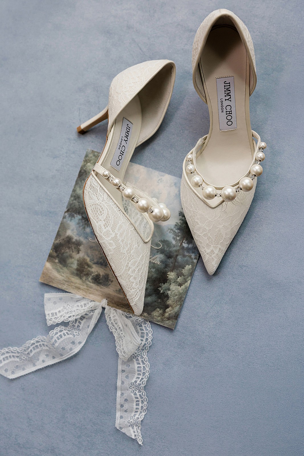 Ivory pointed-toe heels with pearl straps arranged beside lace and a small printed card.
