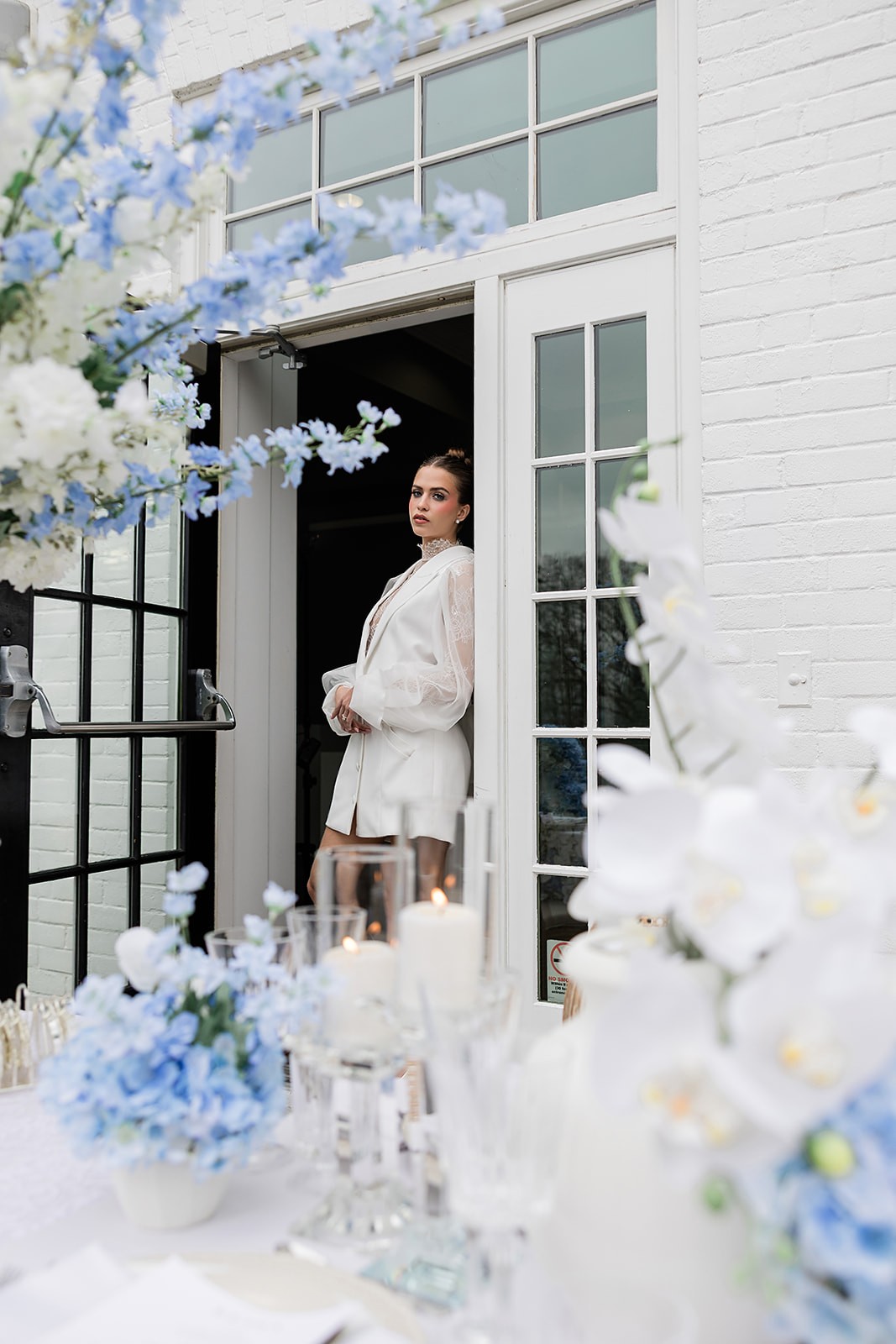 Model in a white blazer dress standing in an open doorway beyond blue-and-white table flowers.