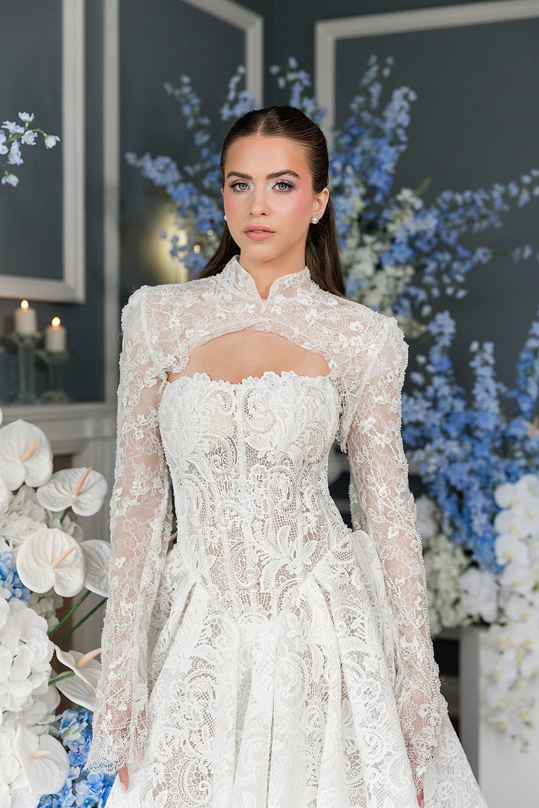 Waist-up portrait of a model in a high-neck lace gown with a keyhole bodice against blue-and-white flowers.