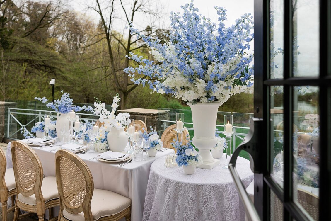 Wide view of a round outdoor table with blue flowers, patterned linen, and cane-back chairs.