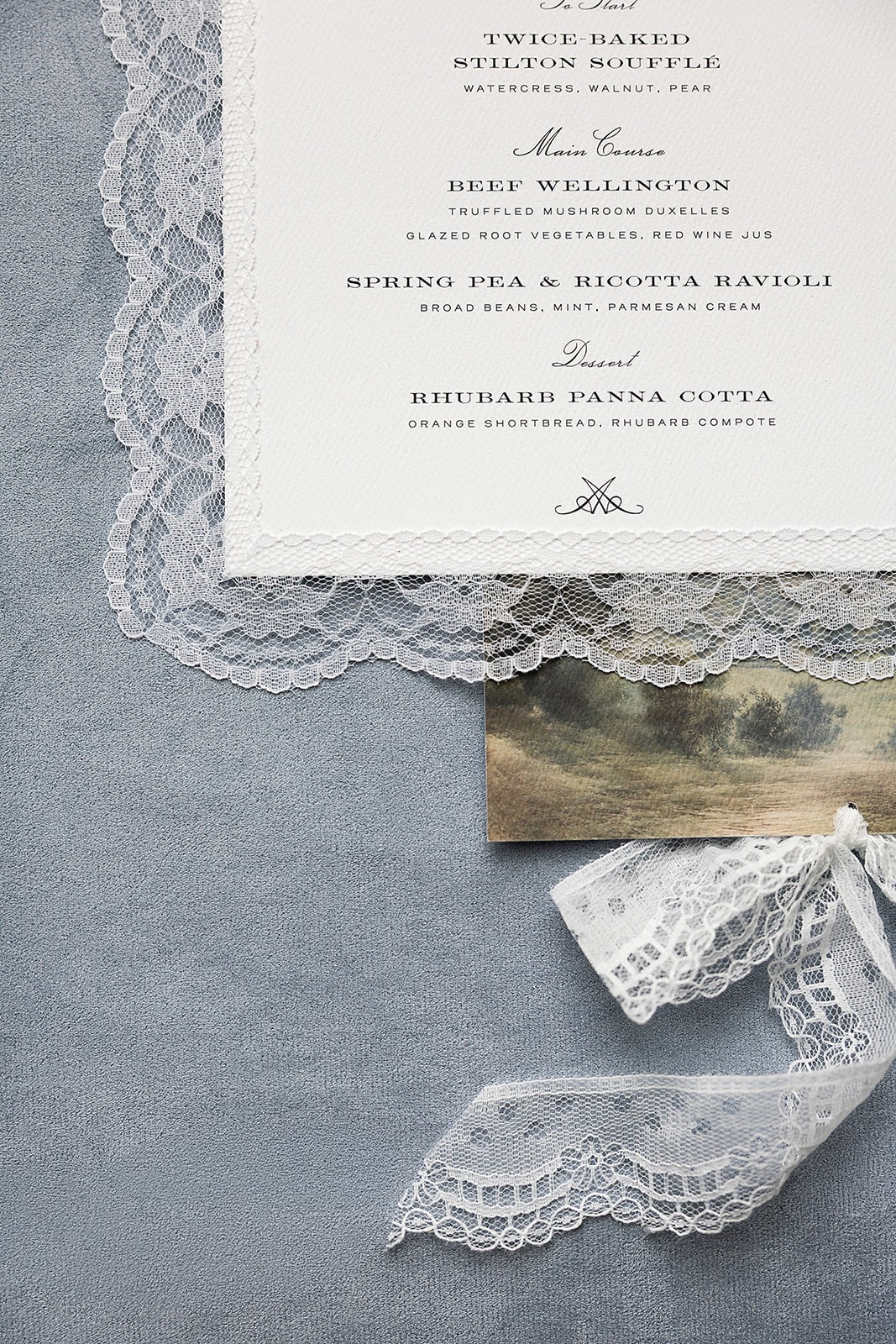 Lace-edged wedding menu card resting on blue fabric beside ribbon and a small printed detail.