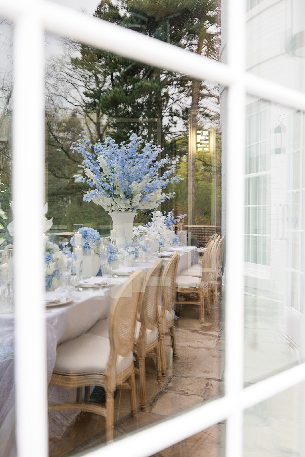 Outdoor reception table seen through white window frames, with blue flowers and cane-back chairs.