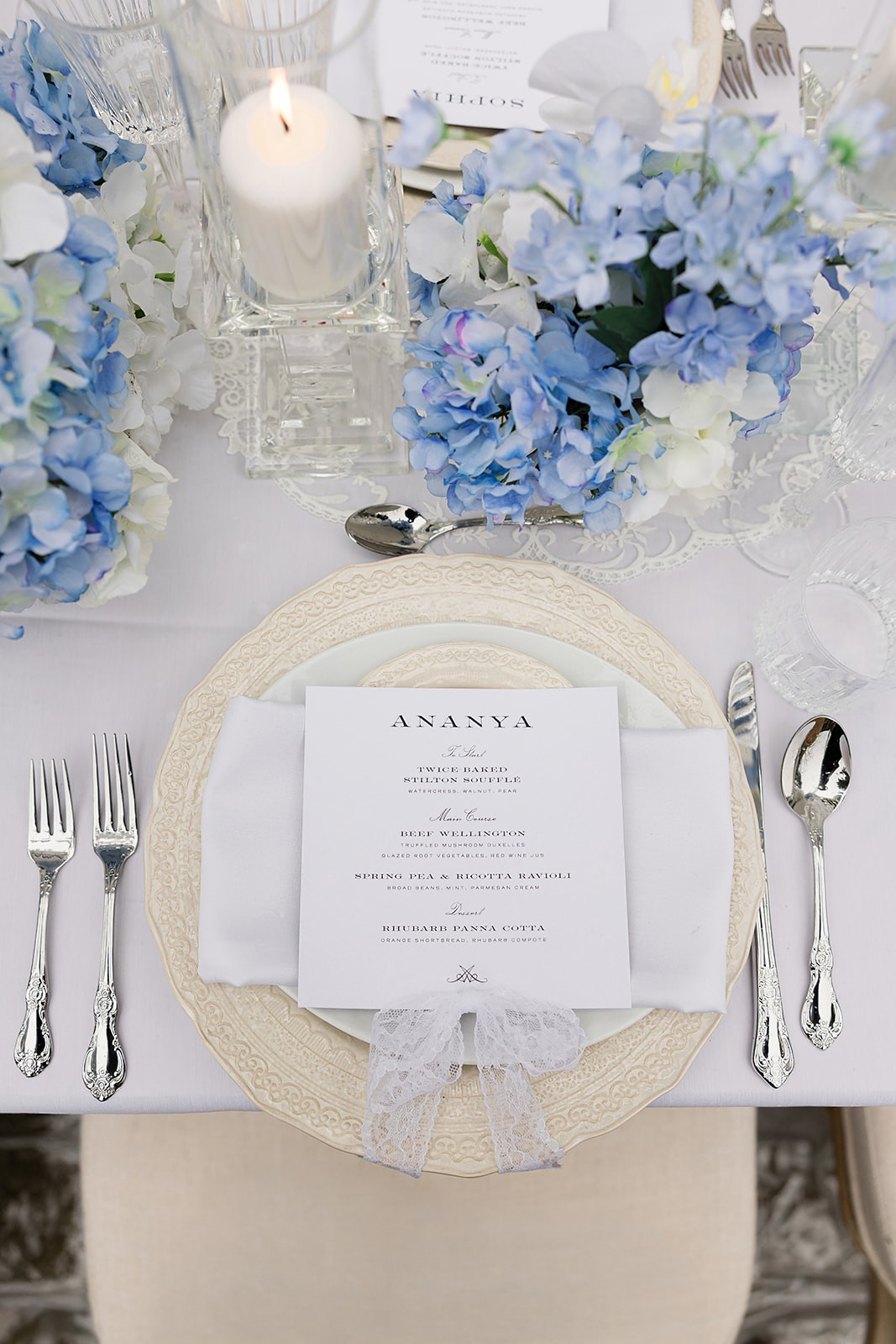 Top-down place setting with a printed menu on layered plates surrounded by pale blue flowers and candles.
