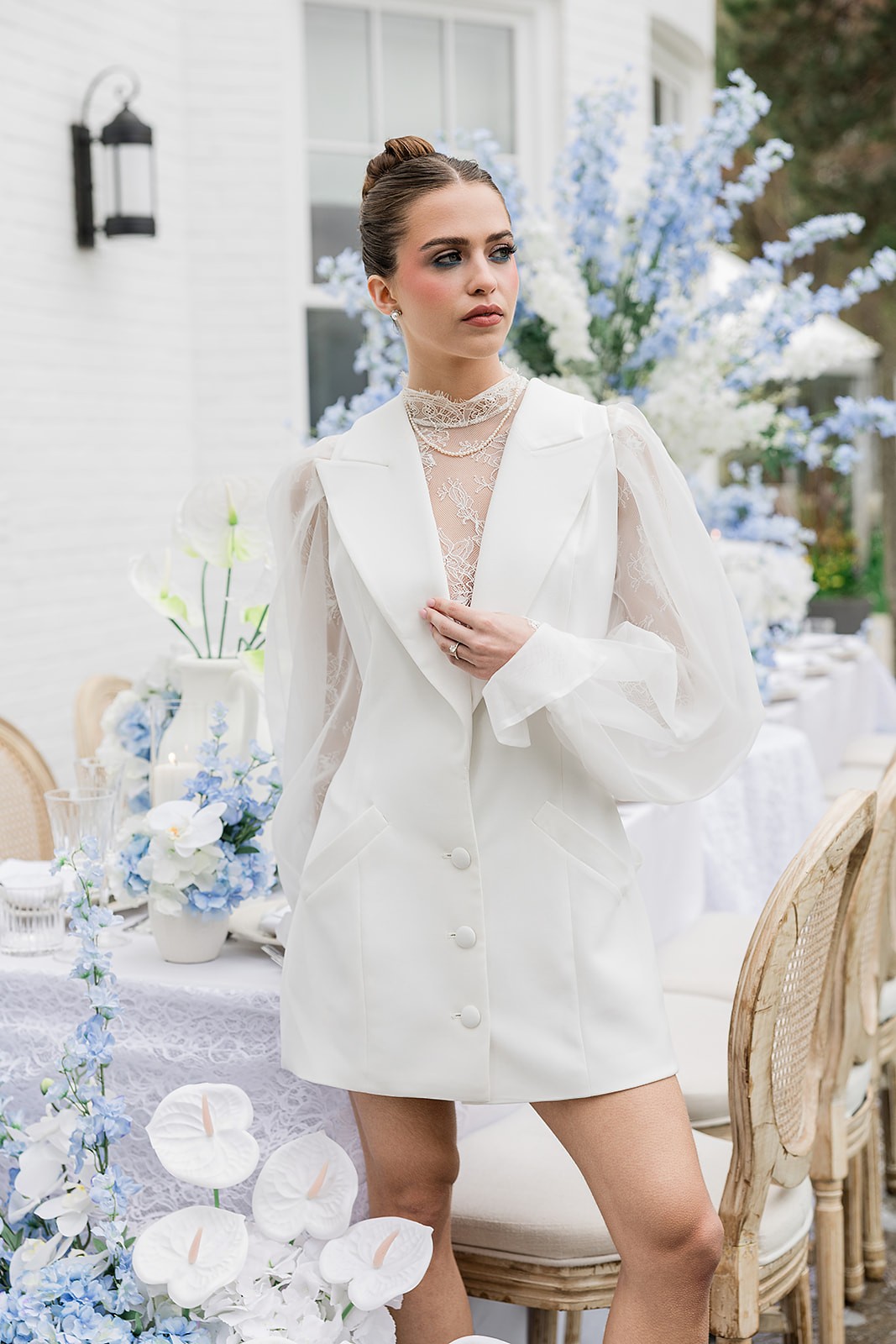 Model in a white blazer dress standing beside an outdoor table and tall blue flowers.