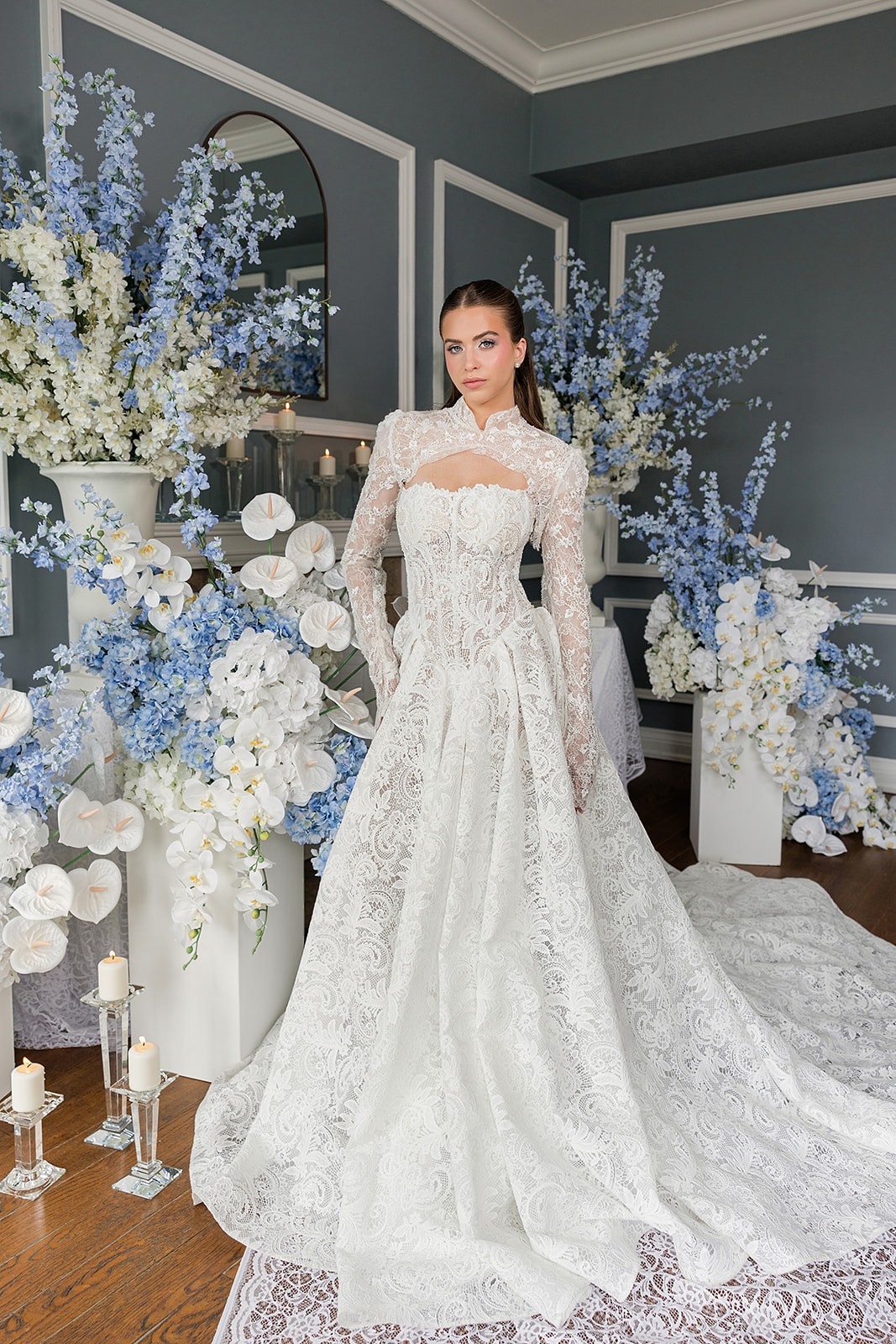 Full-length model in an off-the-shoulder lace ball gown standing before blue-and-white floral installations.