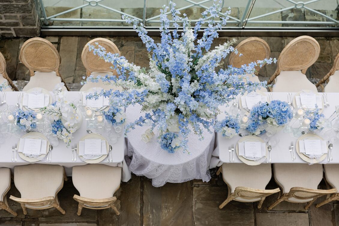 Overhead view of a long outdoor table with blue-and-white flowers, pale place settings, and cane-back chairs.