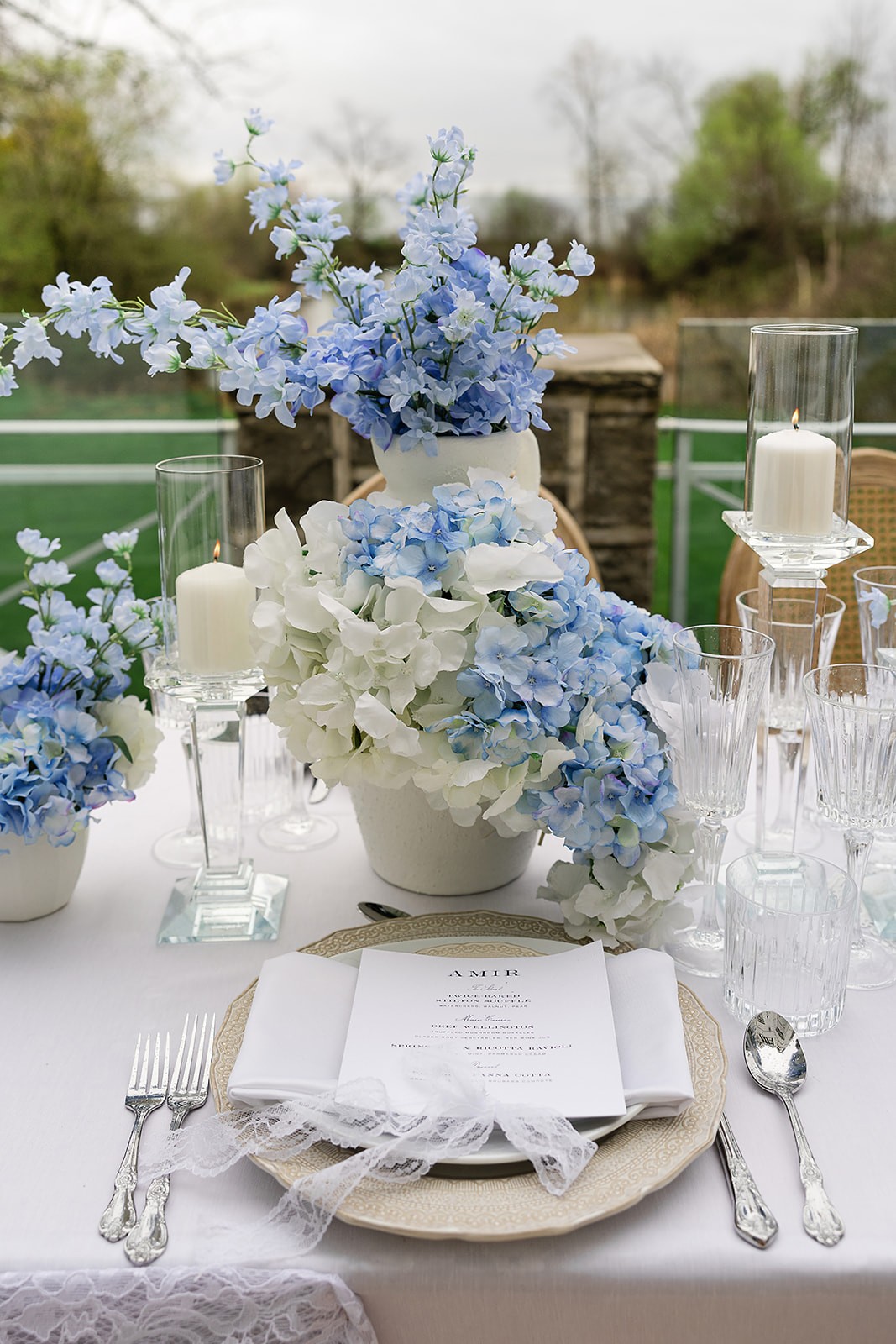 Place setting with a printed menu, silver flatware, glassware, candles, and low blue-and-white flowers.