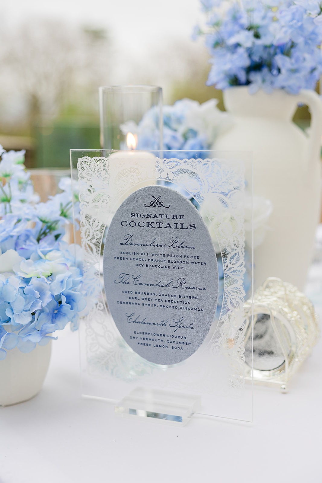 Oval printed cocktail menu standing among pale blue flowers and clear glassware.