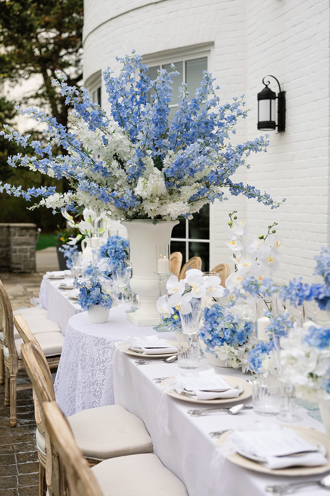 Long outdoor table set with blue-and-white flowers, patterned linen, candles, and pale place settings.