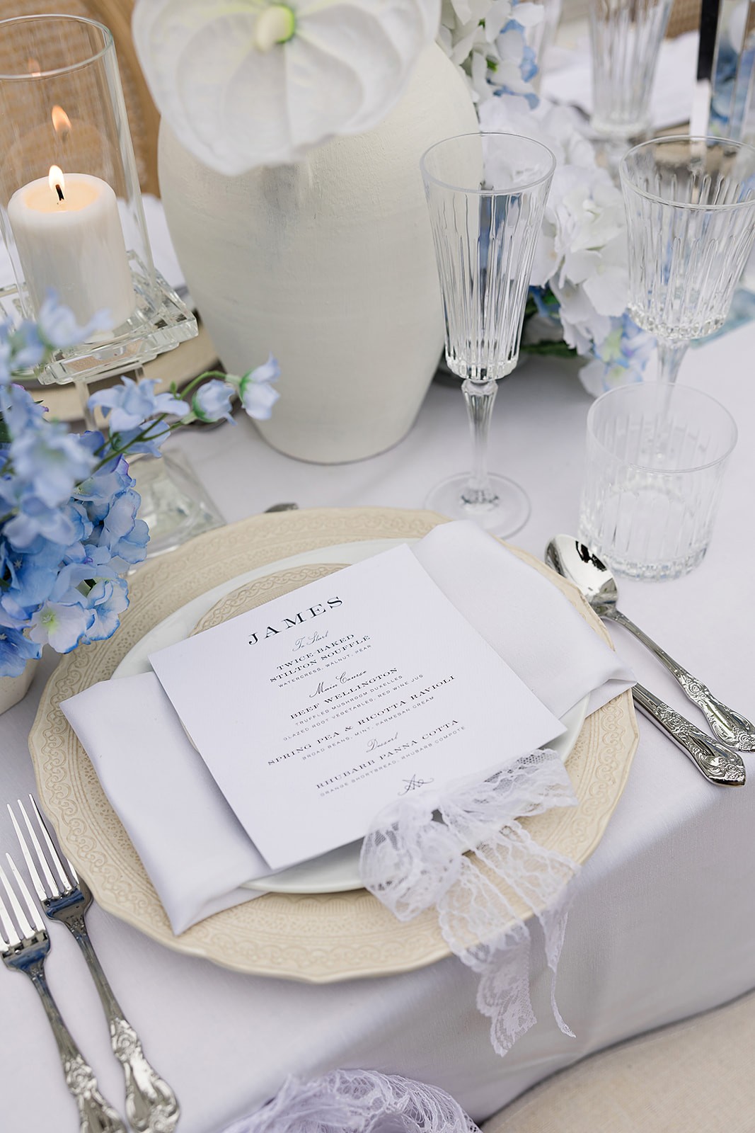 Close view of a printed menu on layered plates beside pale flowers, candles, and glassware.
