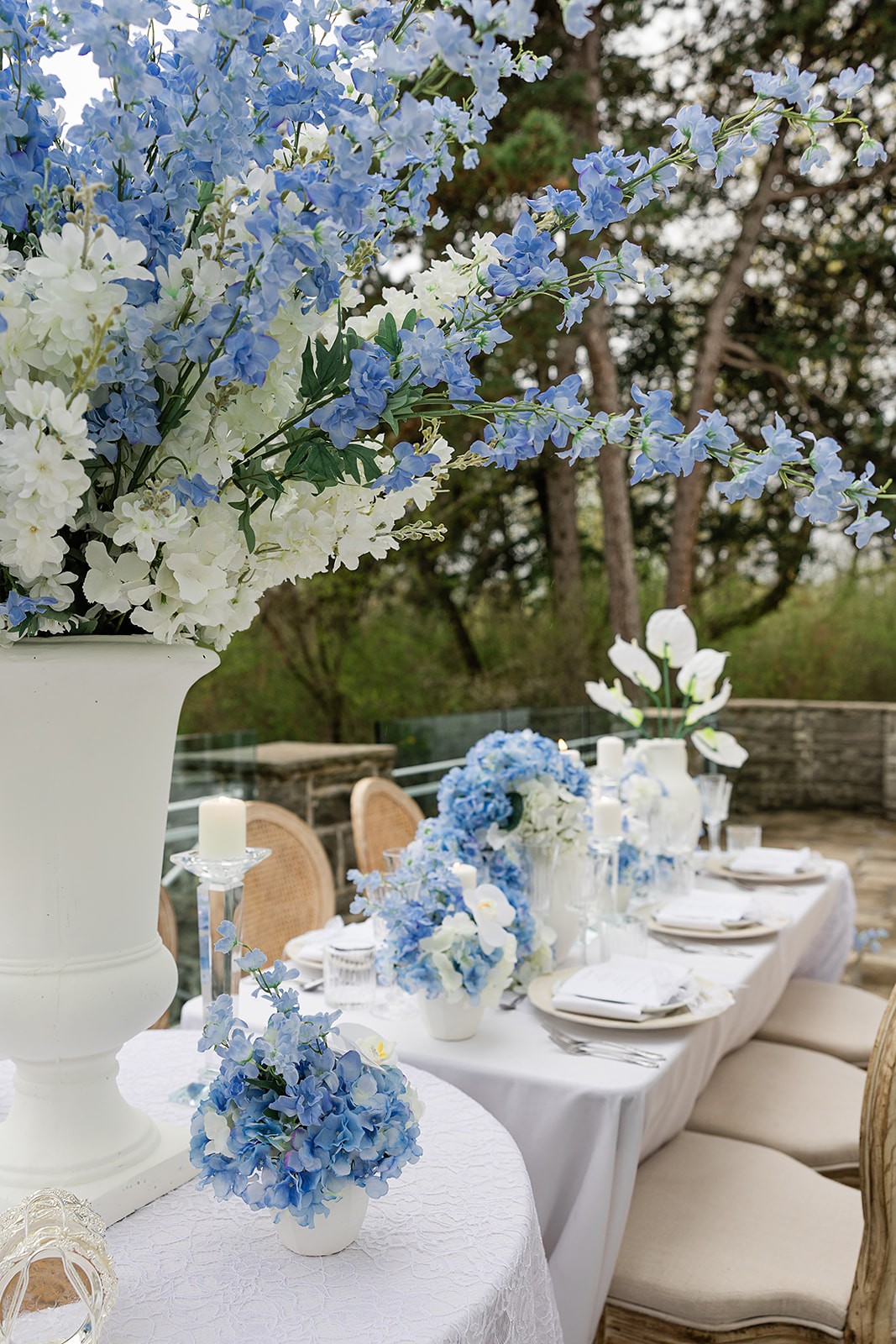 Long outdoor reception table beside the white estate wall with blue flowers and cane-back chairs.