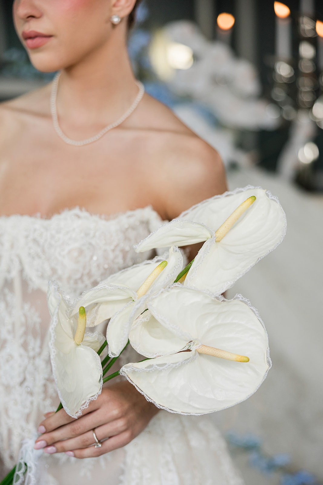Close crop of a strapless lace bodice, pearl necklace, and white anthurium bouquet held at the waist.