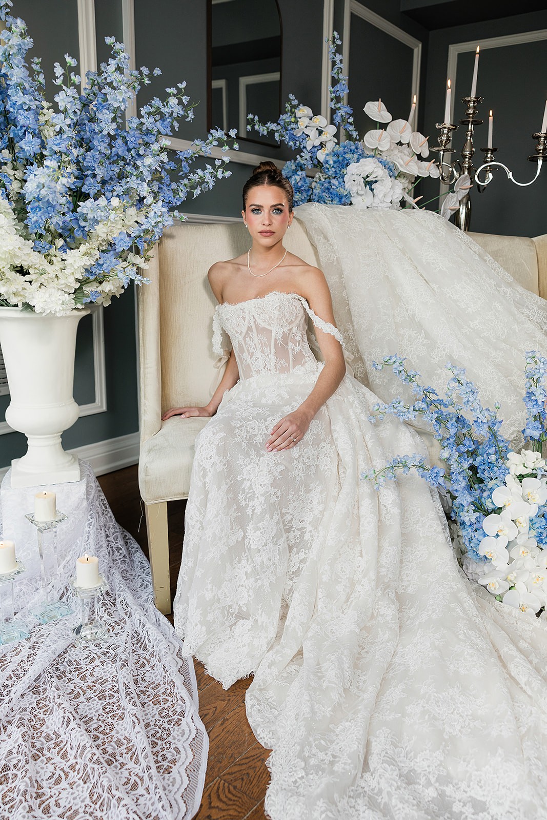 Model seated in a strapless lace ball gown with the skirt spread across the floor among blue-and-white flowers.