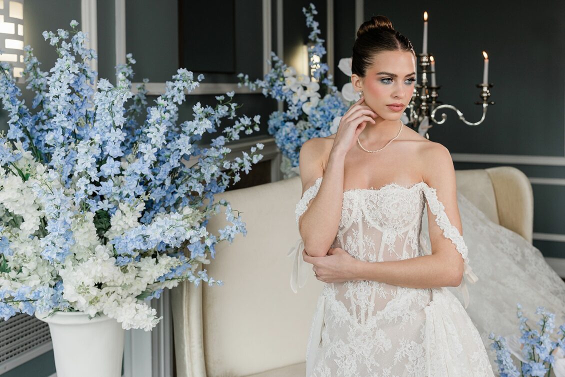 Wide portrait of a model in a strapless lace gown posed beside a pale chair and blue floral branches.