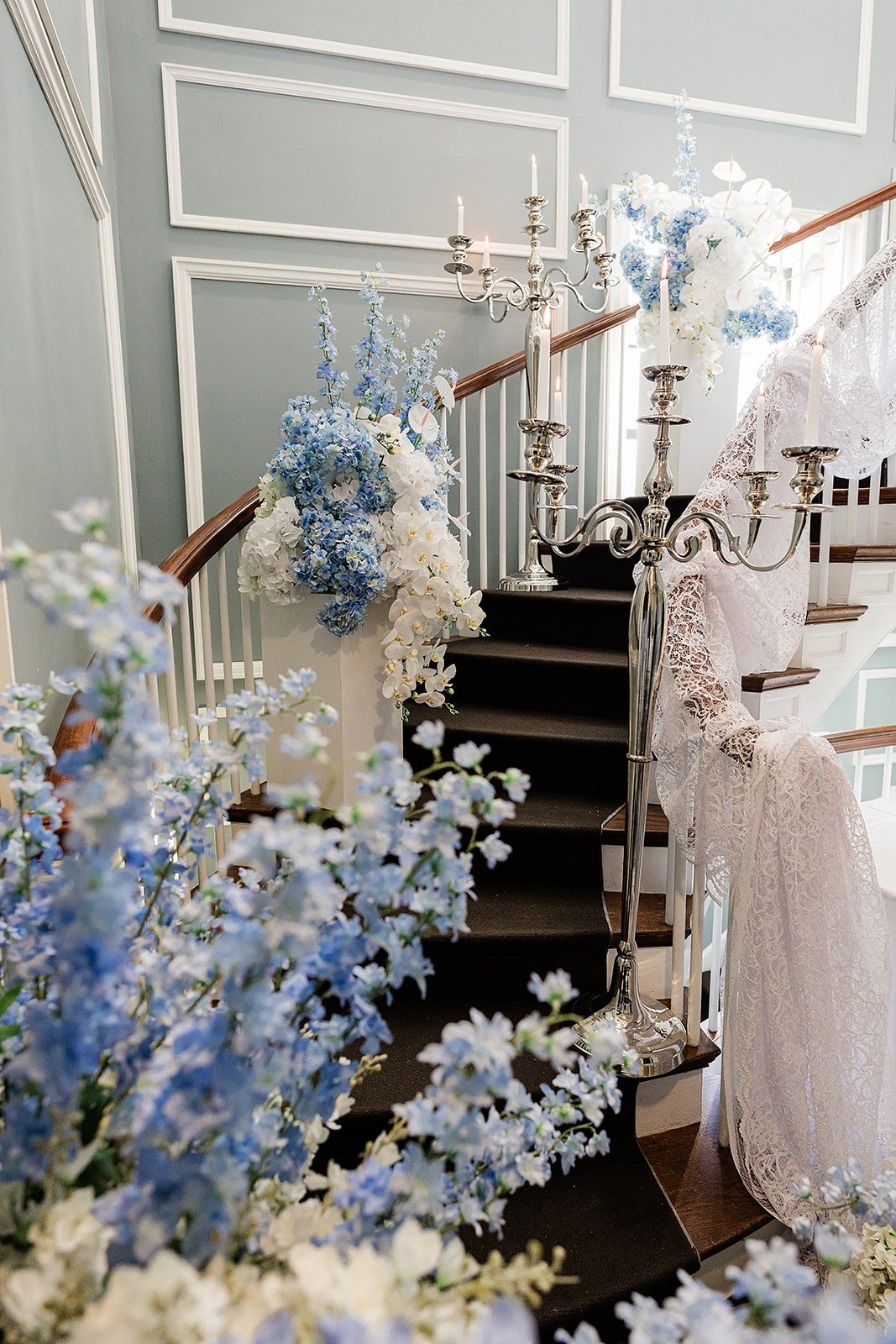Estate staircase decorated with lace, blue-and-white flowers, candles, and an ivory cake on a round table.