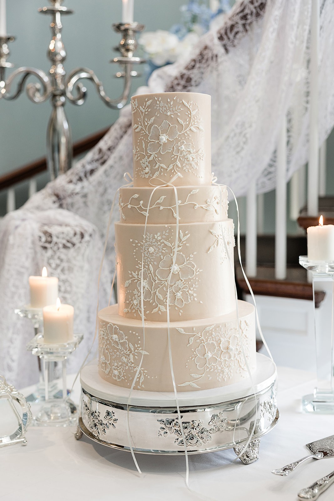 Four-tier ivory wedding cake with delicate white piping displayed beside lace and taper candles.