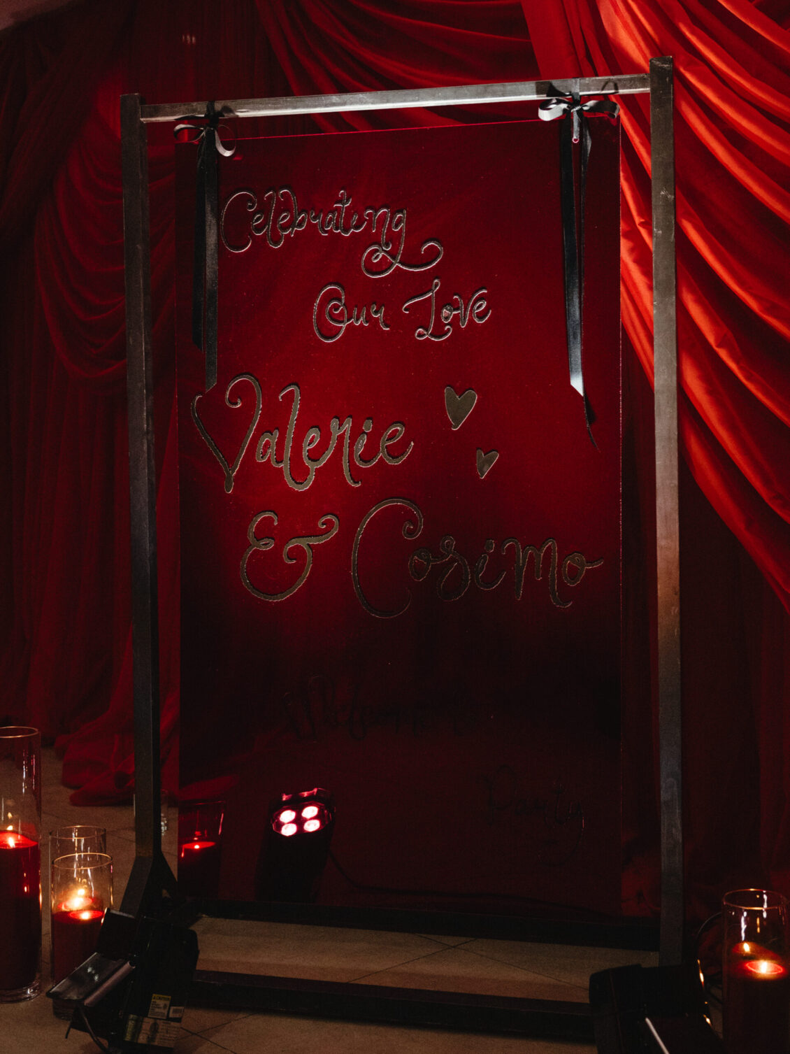 A glass welcome sign with red script and small hearts stands against gathered red drapery and candlelight.