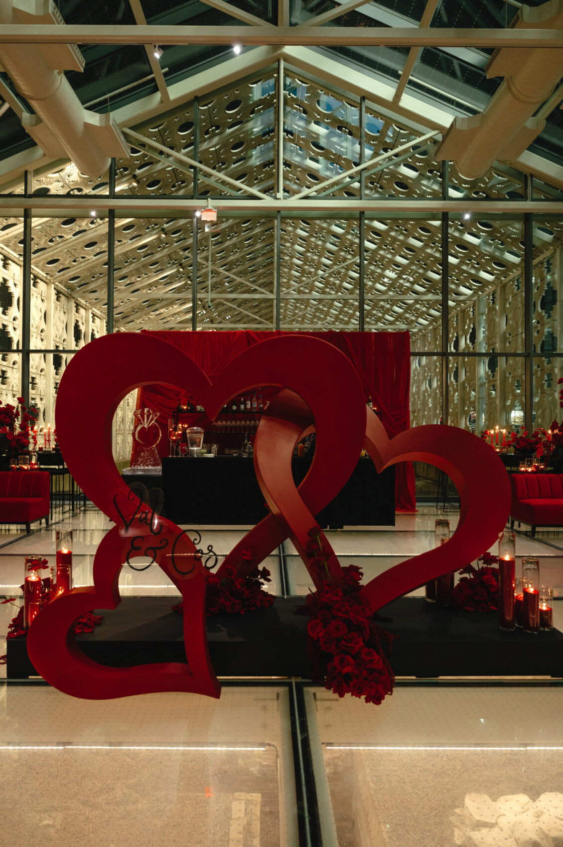 Three intertwined red heart sculptures rise above red flowers and candles beneath a glass roof.