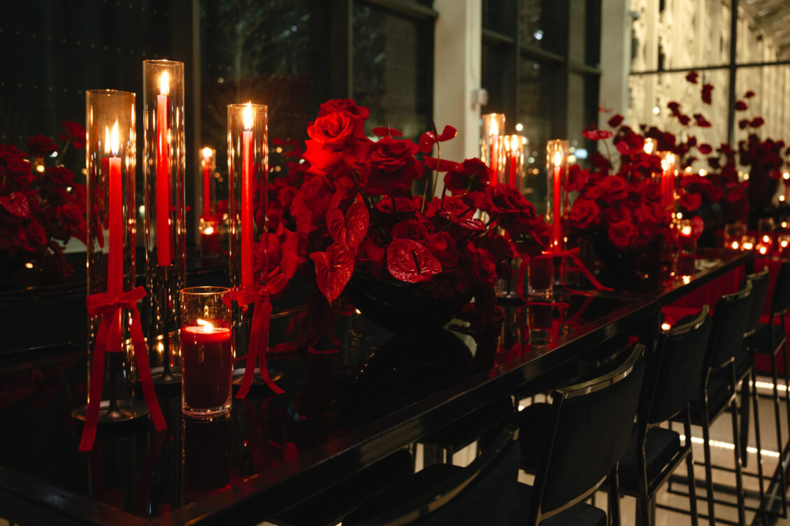 Red roses, red votives, and tall glass candles cover a glossy black dining table lined with black chairs.