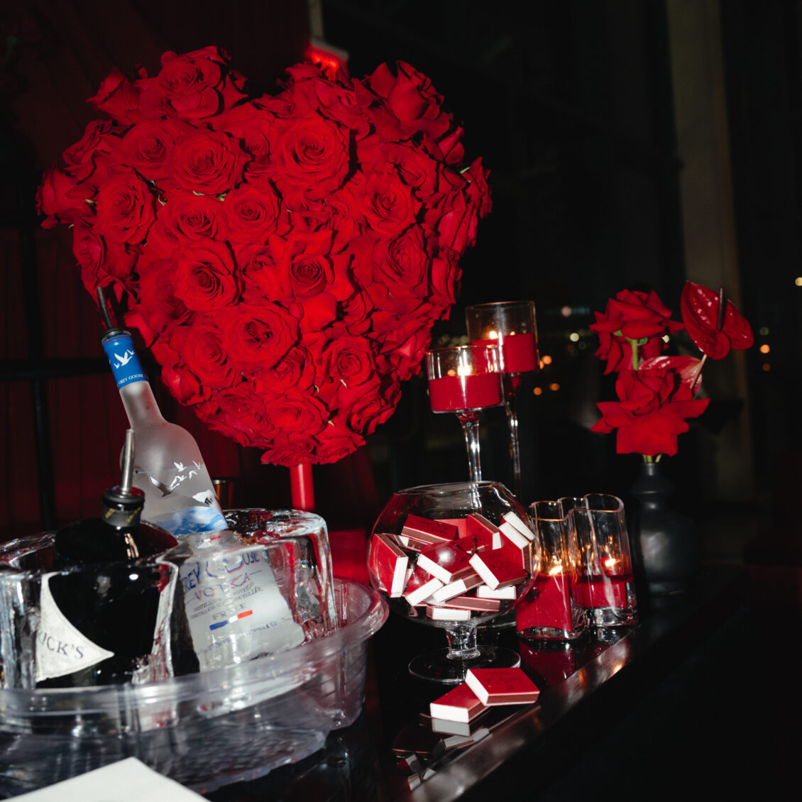 A round bouquet of red roses stands above an ice bucket, glasses, small red-and-white favours, and candles.