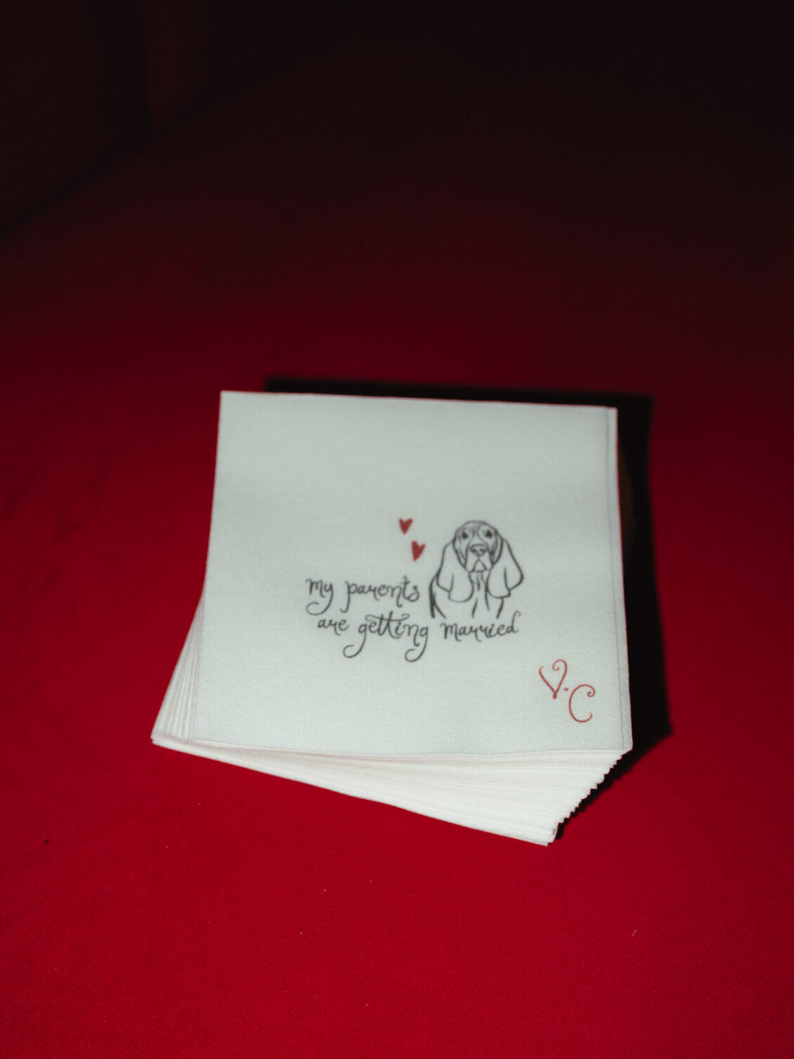 A stack of white cocktail napkins with a couple illustration, red hearts, and red initials rests on a vivid red surface.