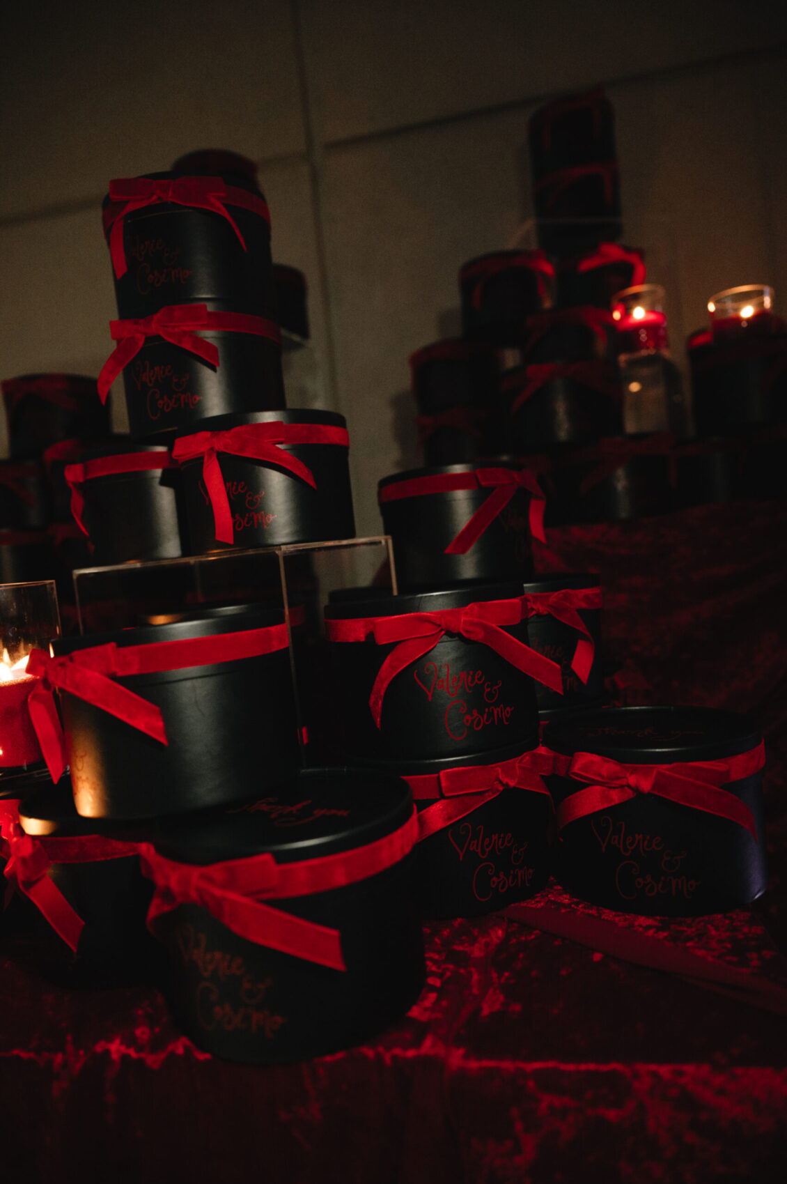 Matte black round favour boxes are stacked with bright red ribbon bows among red petals and candlelight.