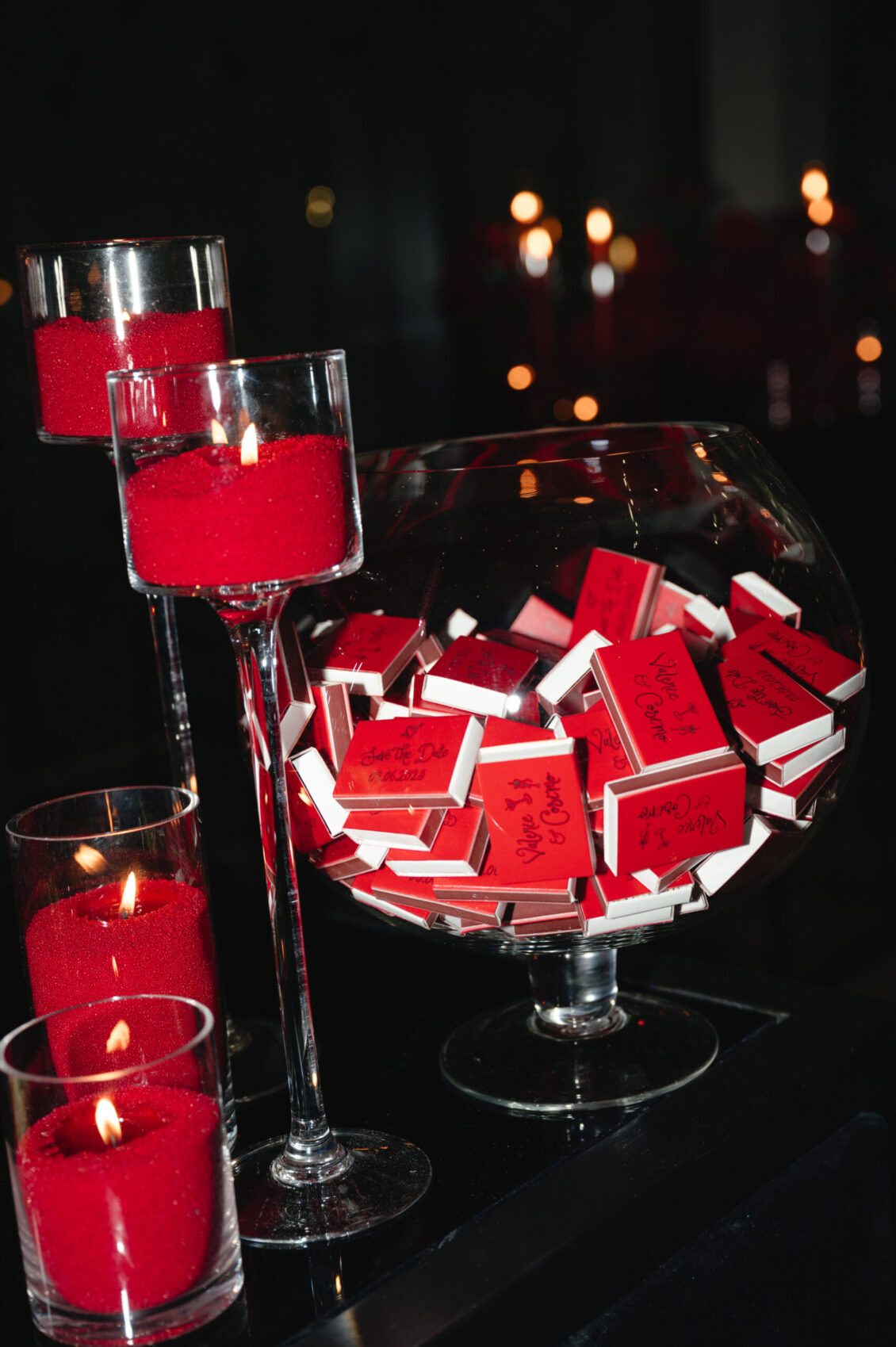 A stemmed glass bowl filled with red-and-white matchbooks sits among red candles on a glossy black surface.