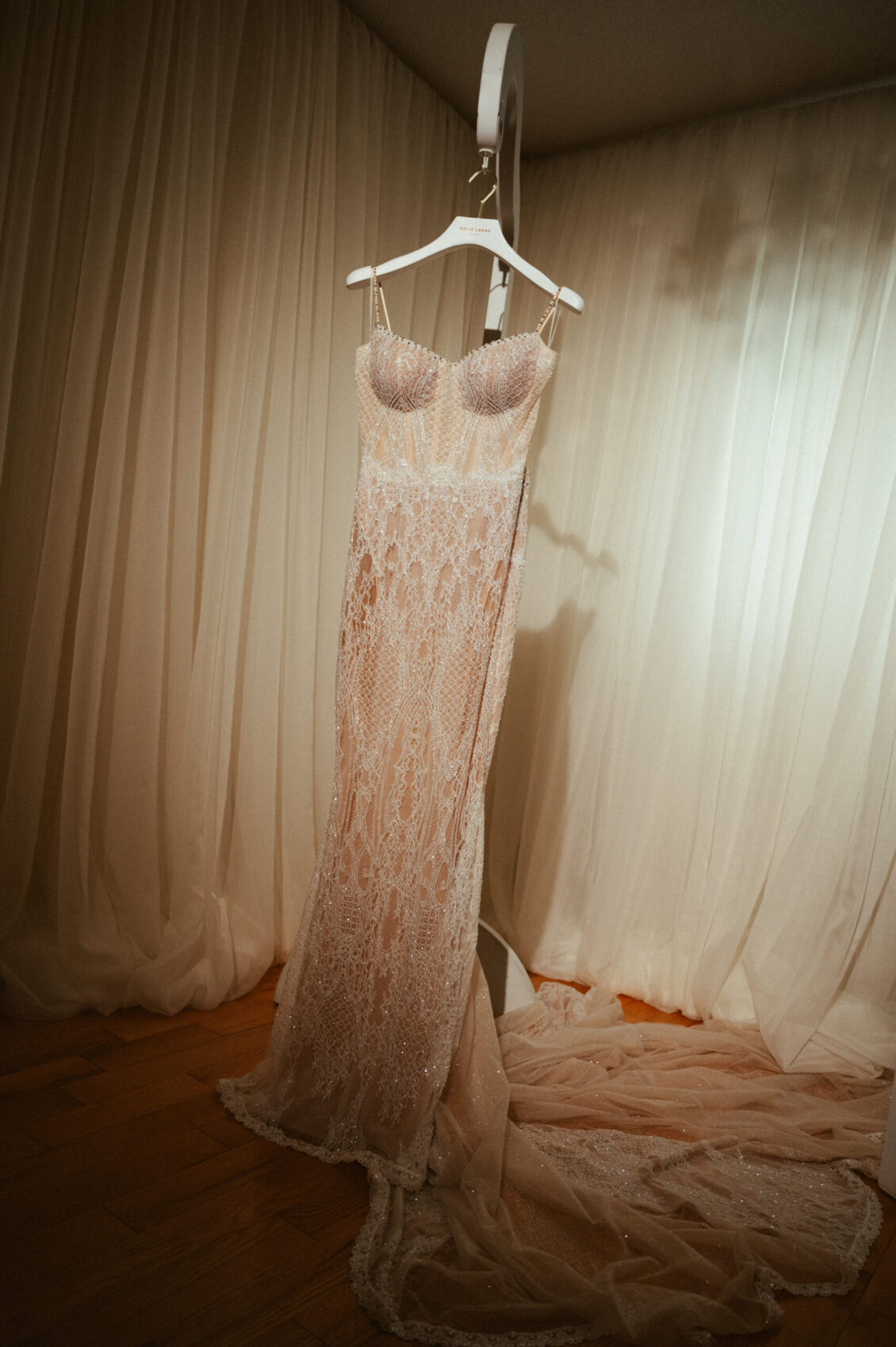 An ivory lace wedding gown hangs before cream curtains with its long train pooled across a wooden floor.