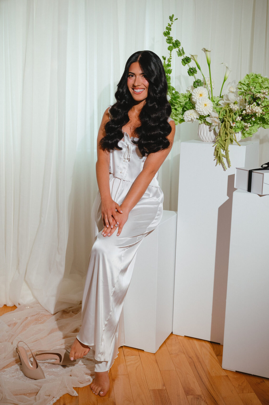 Valerie sits smiling in a white satin robe beside white pedestal blocks and pale floral arrangements.
