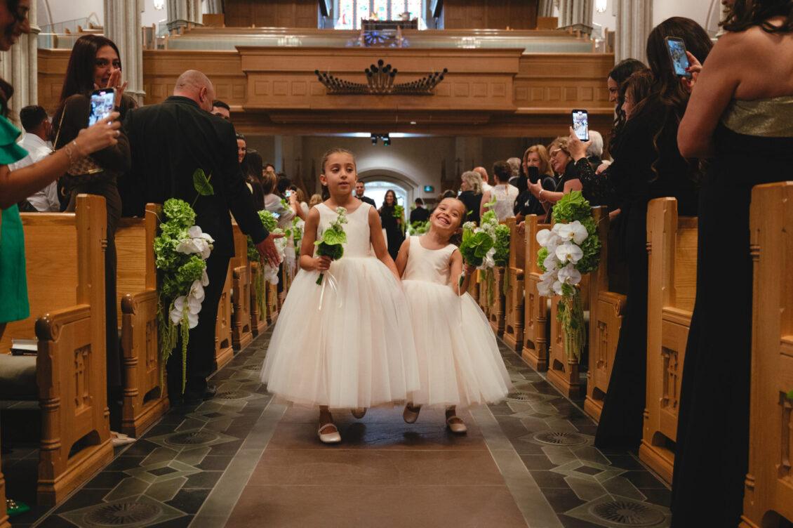 Two young flower girls in long white dresses walk side by side down the cathedral aisle carrying small green-and-white floral pieces.