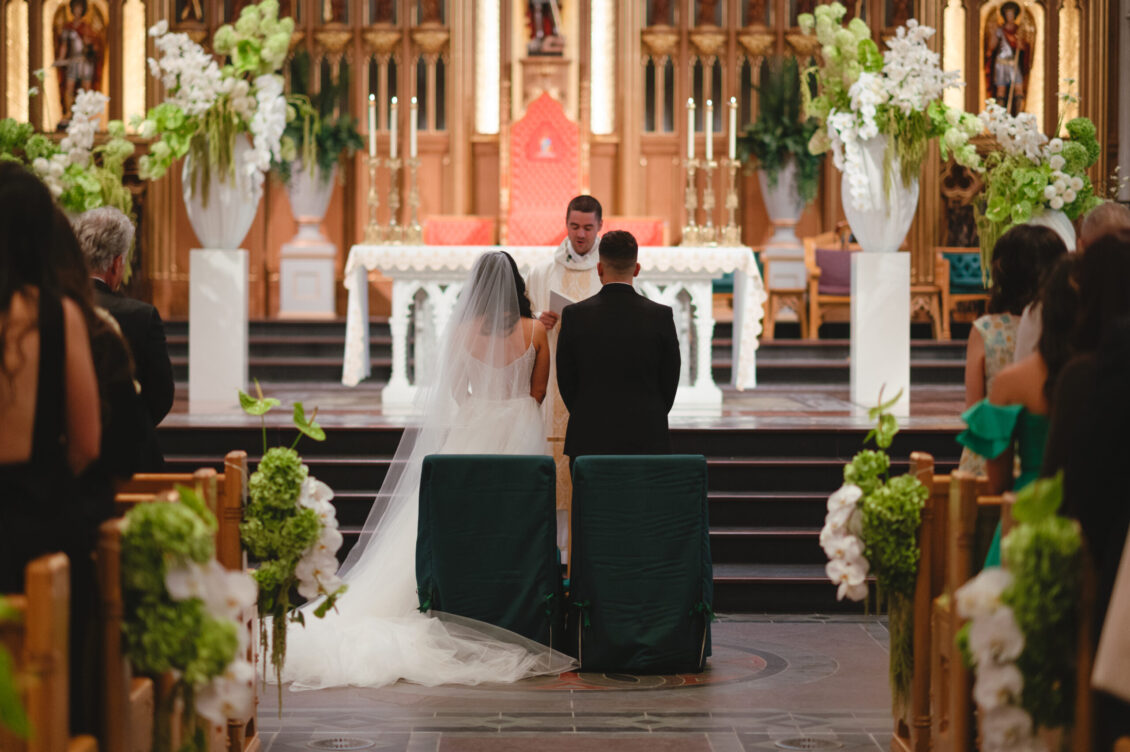 Valerie and Cosimo sit facing a white-robed officiant at the altar amid candles, flowers, statues, and carved woodwork.