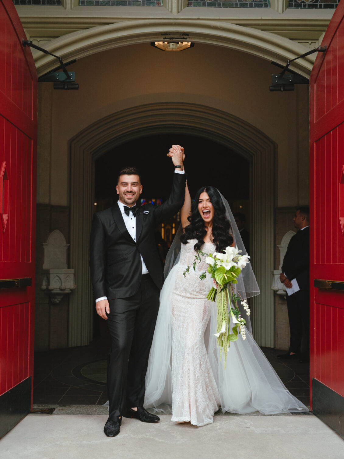 Cosimo raises Valerie's hand as they stand smiling in the open cathedral doorway framed by bright-red doors.