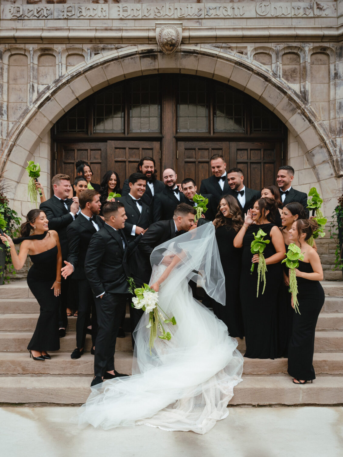 Valerie and Cosimo pose with a large cheering wedding party on stone church steps beneath an arched wooden doorway.