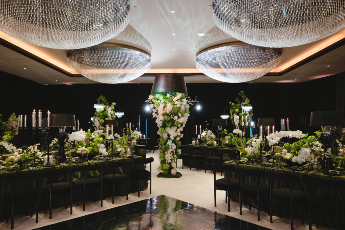 Long black tables, dark chairs, white floral installations, and rounded crystal chandeliers surround a glossy reception aisle.