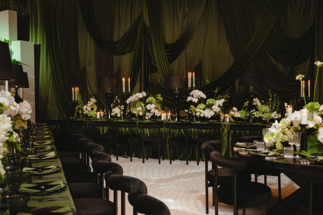 Long olive-green tables, black chairs, white flowers, candles, black lamps, and gathered green drapery fill an empty reception room.