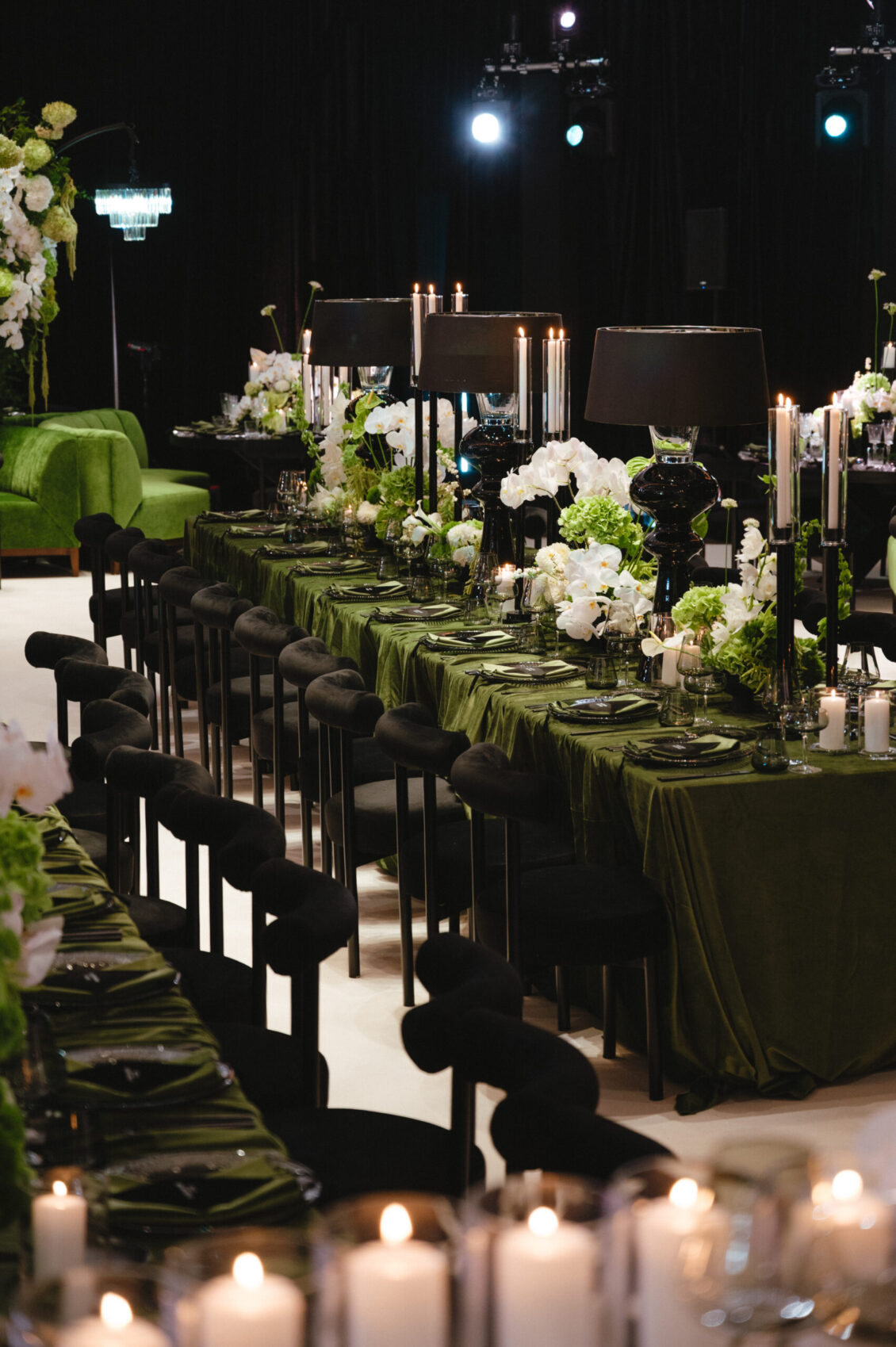 A long olive-green banquet table is lined with black chairs, glassware, white flowers, candles, and glowing votives.