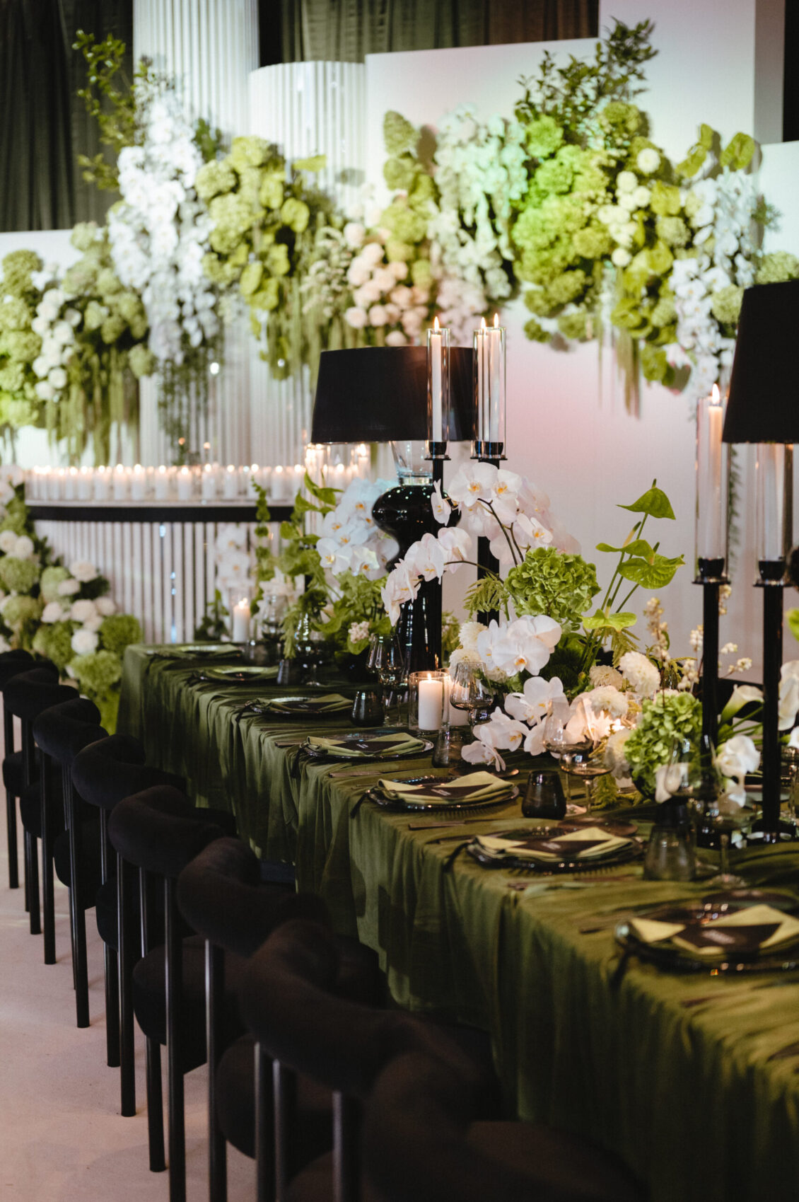An olive-green table with black chairs, flowers, candles, and black lamps stands before a wall of hanging pale blooms.