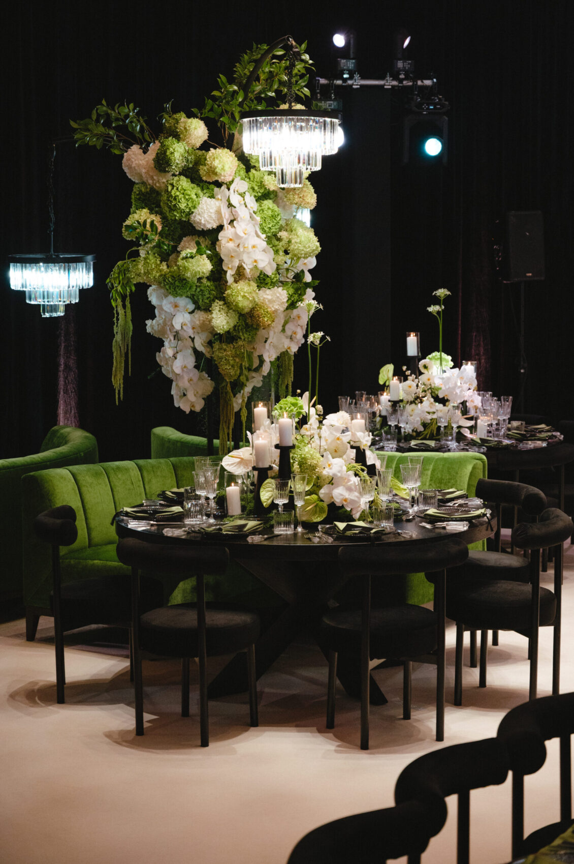 A tall cascading white, blush, and green floral arrangement rises above a candlelit round table surrounded by black and green seating.