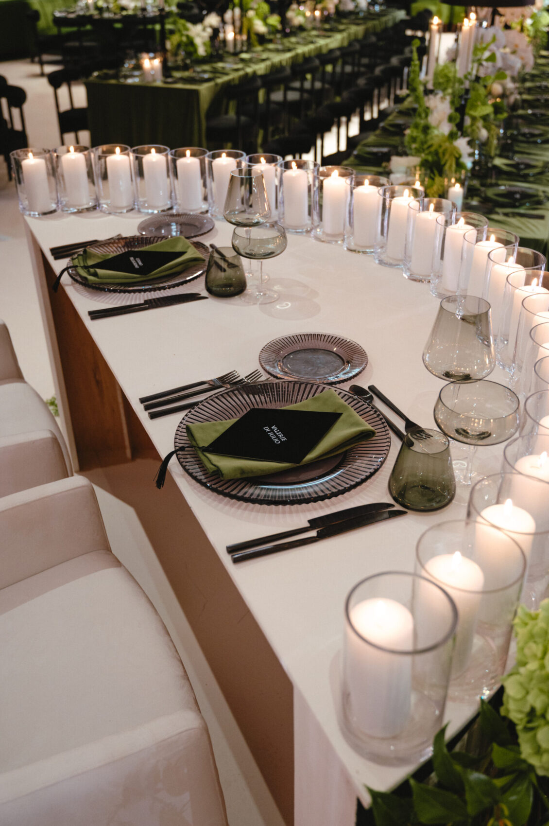 Clear cylindrical candles line a white table set with striped glass chargers, dark plates, green napkins, black cards, and black cutlery.