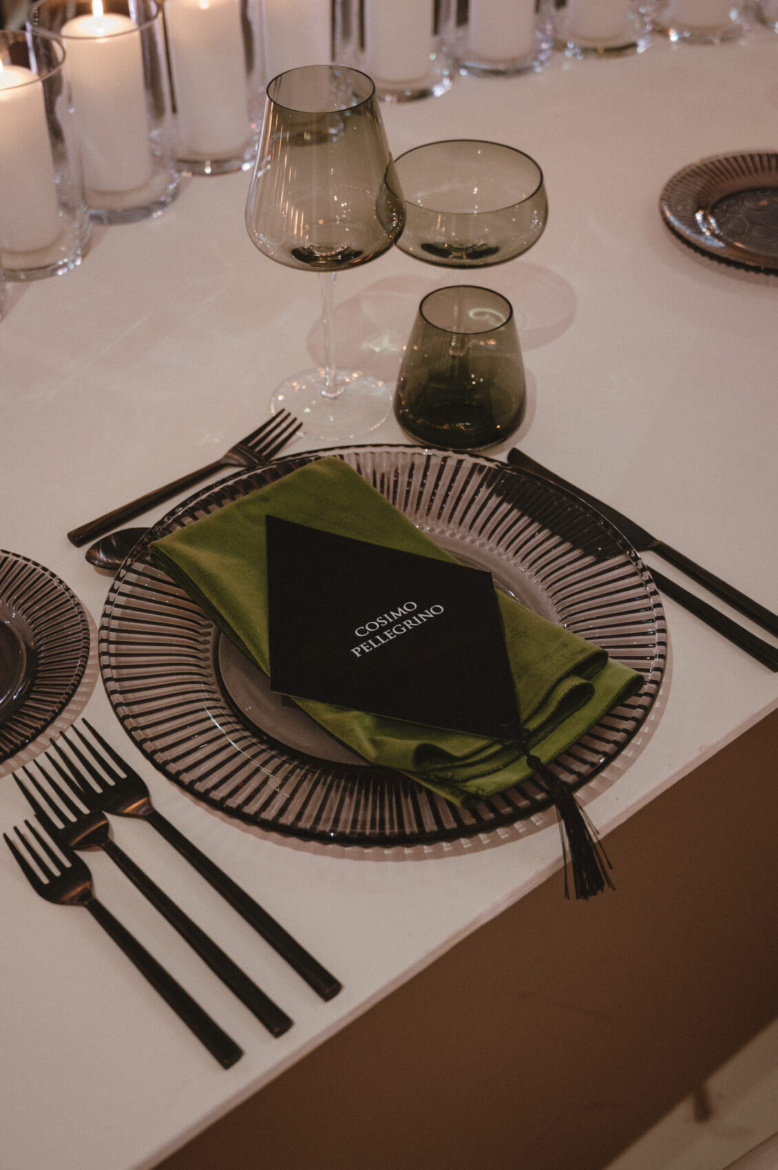 A ribbed glass charger holds a dark plate, folded olive napkin, and black card beside black flatware and smoky glasses.