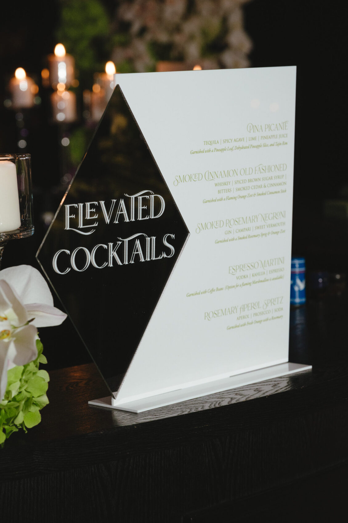 A glossy black Elevated Cocktails sign and tall printed menu stand beside white flowers and warm candles on a black table.