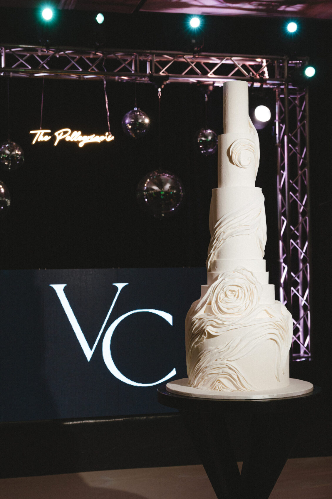 A tall white floral-relief wedding cake stands on a black pedestal before large white VC letters and disco balls.